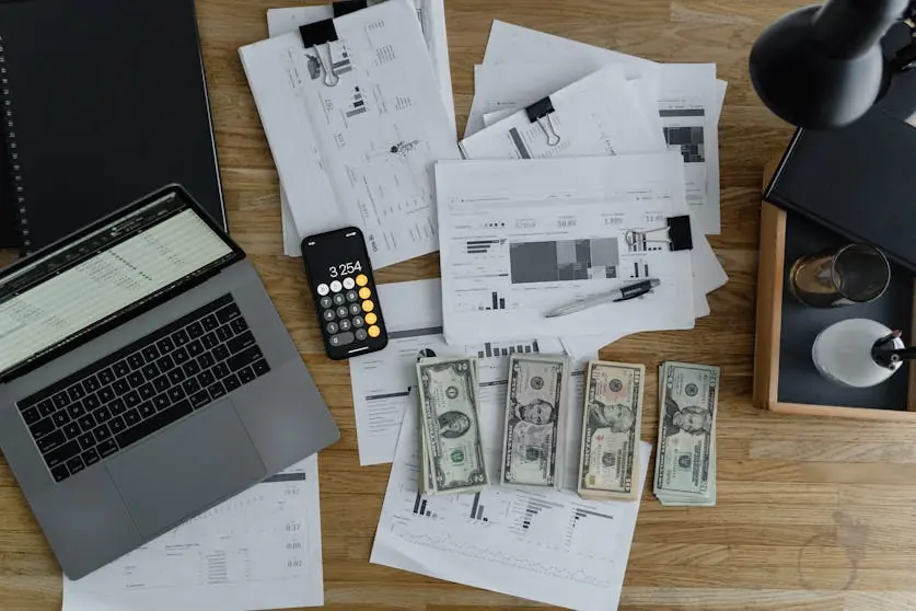 Una scrivania con sopra un portatile, dei documenti finanziari e contanti.